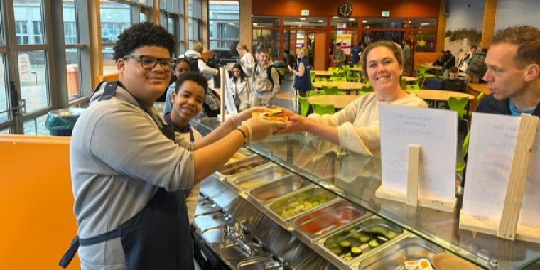 Twee cafetariamedewerkers glimlachen en geven een bord eten aan een vrouw en een man aan de andere kant van de toonbank. Mensen zitten en lopen in de lichte, drukke cafetaria op de achtergrond.
