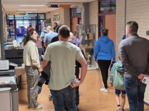 Een groep volwassenen en kinderen loopt door een schoolgang met beige bakstenen muren, glazen vitrines en klaslokalen. Sommige mensen lopen samen, anderen kijken om zich heen. De vloer is bruin en het gebied is fel verlicht.