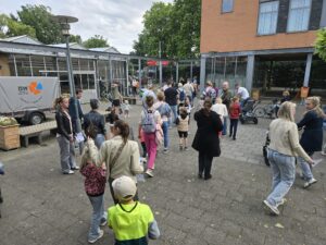 Een groep volwassenen en kinderen verzamelt zich voor een modern gebouw met grote ramen. Sommigen houden papieren vast en een paar kinderen hebben rugzakken. Bomen en een geparkeerd busje zijn op de achtergrond zichtbaar.