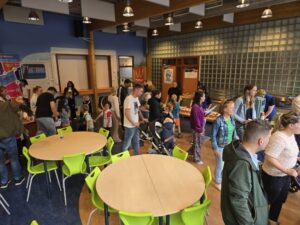 Een groep mensen, bestaande uit volwassenen en kinderen, verzamelt zich in een felverlichte binnenruimte met ronde tafels en groene stoelen. Sommigen staan in de rij, terwijl anderen praten of naar tafels met voorwerpen kijken.
