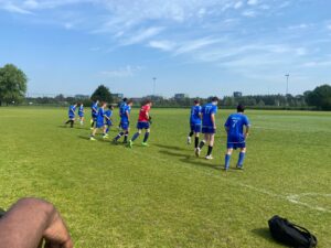 Een groep voetballers in blauwe tenues jogt samen op een grasveld onder een strakblauwe lucht, om zich op te warmen voor een wedstrijd. Eén speler draagt een rood shirt en sporttassen liggen op het grasveld in de buurt. Bomen en gebouwen zijn op de achtergrond te zien.