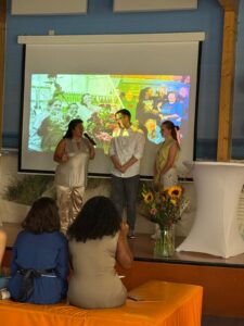Drie mensen staan te praten op een klein podium met een boeket zonnebloemen ervoor. Twee mensen zitten op de voorgrond. Achter de sprekers wordt een kleurrijk geprojecteerd beeld met lachende gezichten getoond.