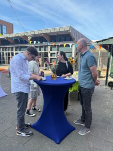 Vier mensen staan buiten rond een hoge ronde tafel, gedekt met een blauw kleed, te praten en te schrijven. Erboven hangen kleurrijke slingers en de tafel is versierd met bloemen. Het is zonnig weer met een heldere hemel.
