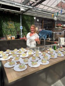 Een glimlachende vrouw staat achter een toonbank gevuld met vele borden met gelaagde cakeplakken, elk gegarneerd met fruit en slagroom, in een lichte, moderne keuken met planten en een neon koffiebord op de achtergrond.