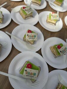 Verschillende witte papieren bordjes, elk met een plak cake in laagjes bedekt met groen glazuur, een kleine rode versiering en een kaartje Geslaagd gefeliciteerd, staan op een houten tafel. Elk bord heeft een wit plastic vorkje.