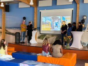 Een groep mensen staat op een podium voor een scherm waarop gymactiviteiten te zien zijn, terwijl een publiek van drie vrouwen op kleurrijke kussens toekijkt. De zaal heeft een strandthema en zonnebloemen op een hoge tafel.