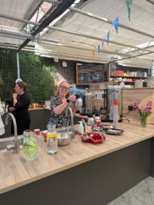 Een glimlachende vrouw met bril staat achter een toonbank met drankjes en snacks en steekt haar duim op. Een andere vrouw staat vlakbij onder een afdak, met planten en kleurrijke gors op de achtergrond.
