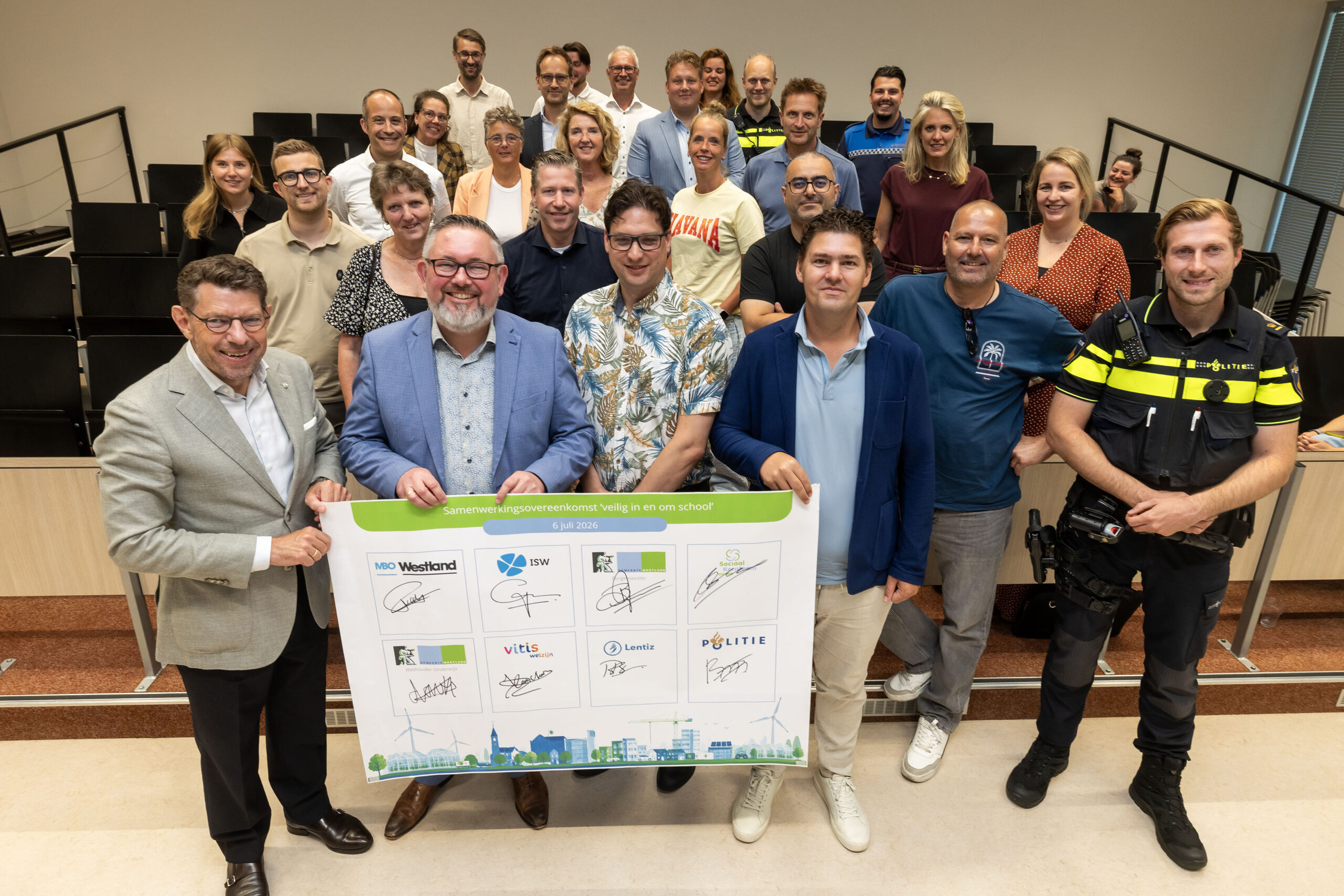 Een groep van ongeveer 25 mensen, waaronder professionals en een politieagent, staat bij elkaar in een klaslokaal met een groot spandoek waarop de ondertekende overeenkomst staat afgebeeld, en lacht naar de camera.