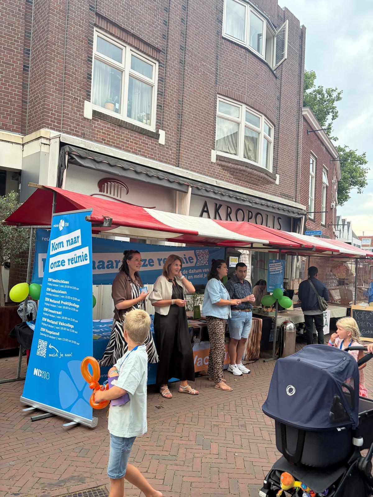 Voor een bakstenen gebouw staan mensen bij een kraampje met een blauw bord en ballonnen. Op de voorgrond staan kinderen met ballondieren en een kinderwagen. Op de tafel van het kraampje liggen posters en folders.
