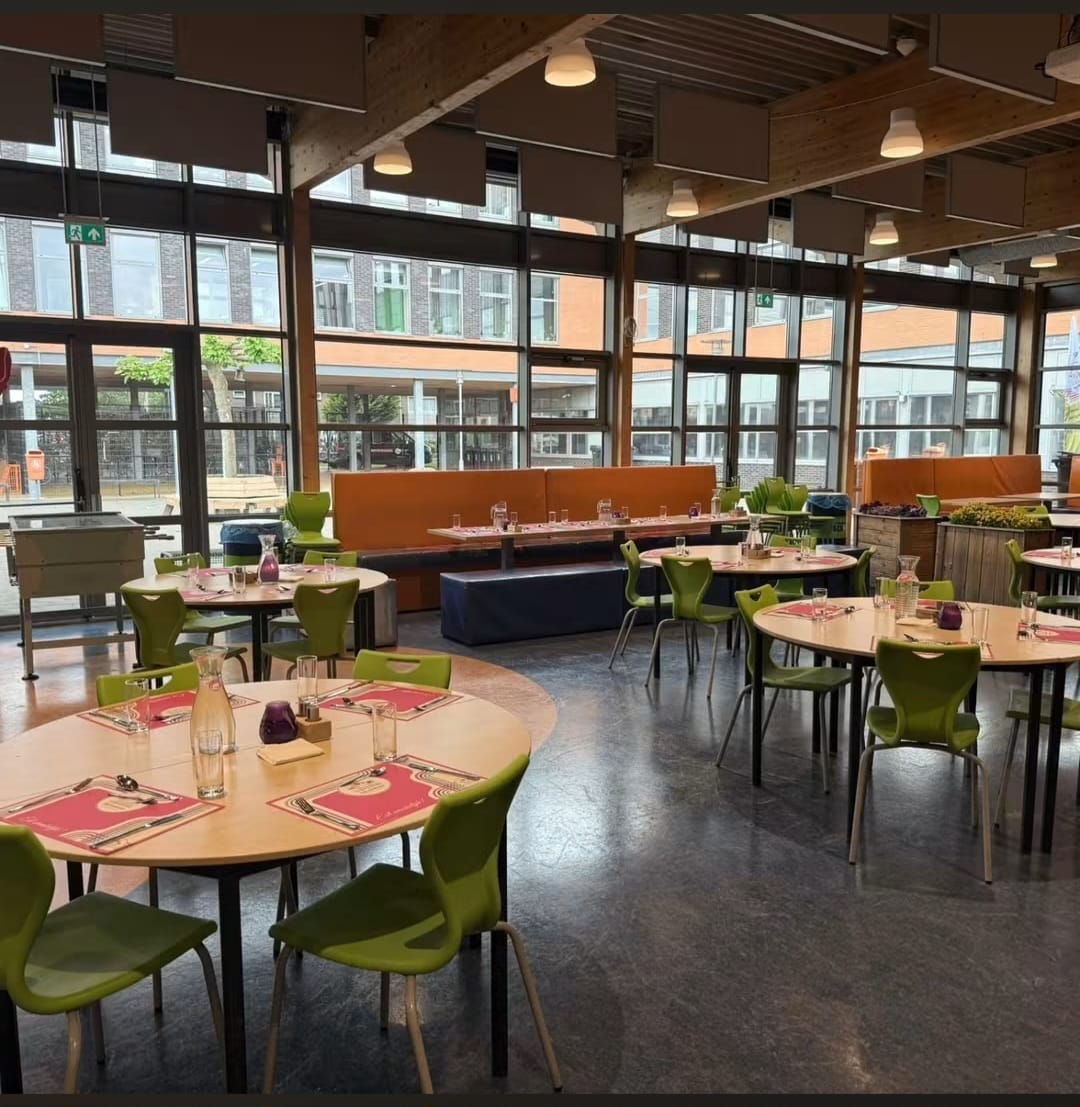 Eetkamer met ronde tafels, groene stoelen en oranje zitbanken, gelegen in een lichte ruimte met ramen van vloer tot plafond.